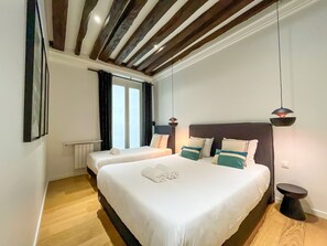 A hotel room with two beds, wooden flooring, a modern ceiling, and a window with curtains.