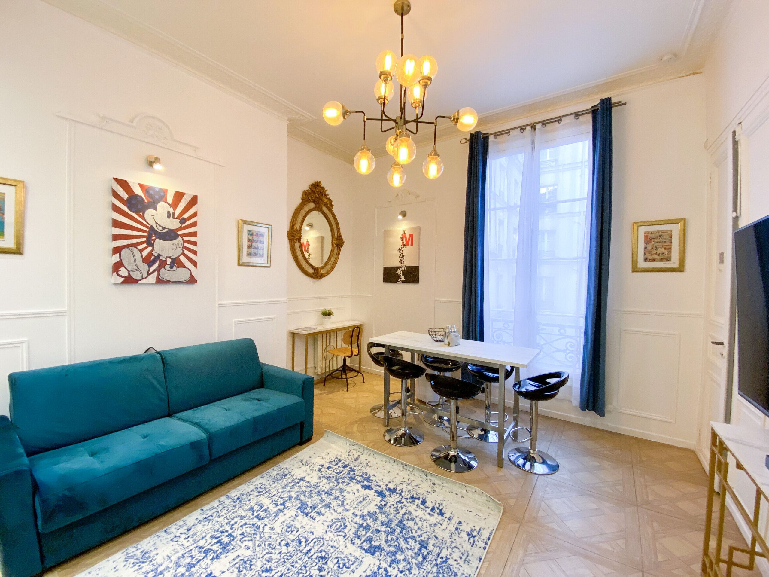 A living room with a teal sofa, a dining area with a white table and four black bar stools, a chandelier, and a large window with blue curtains.