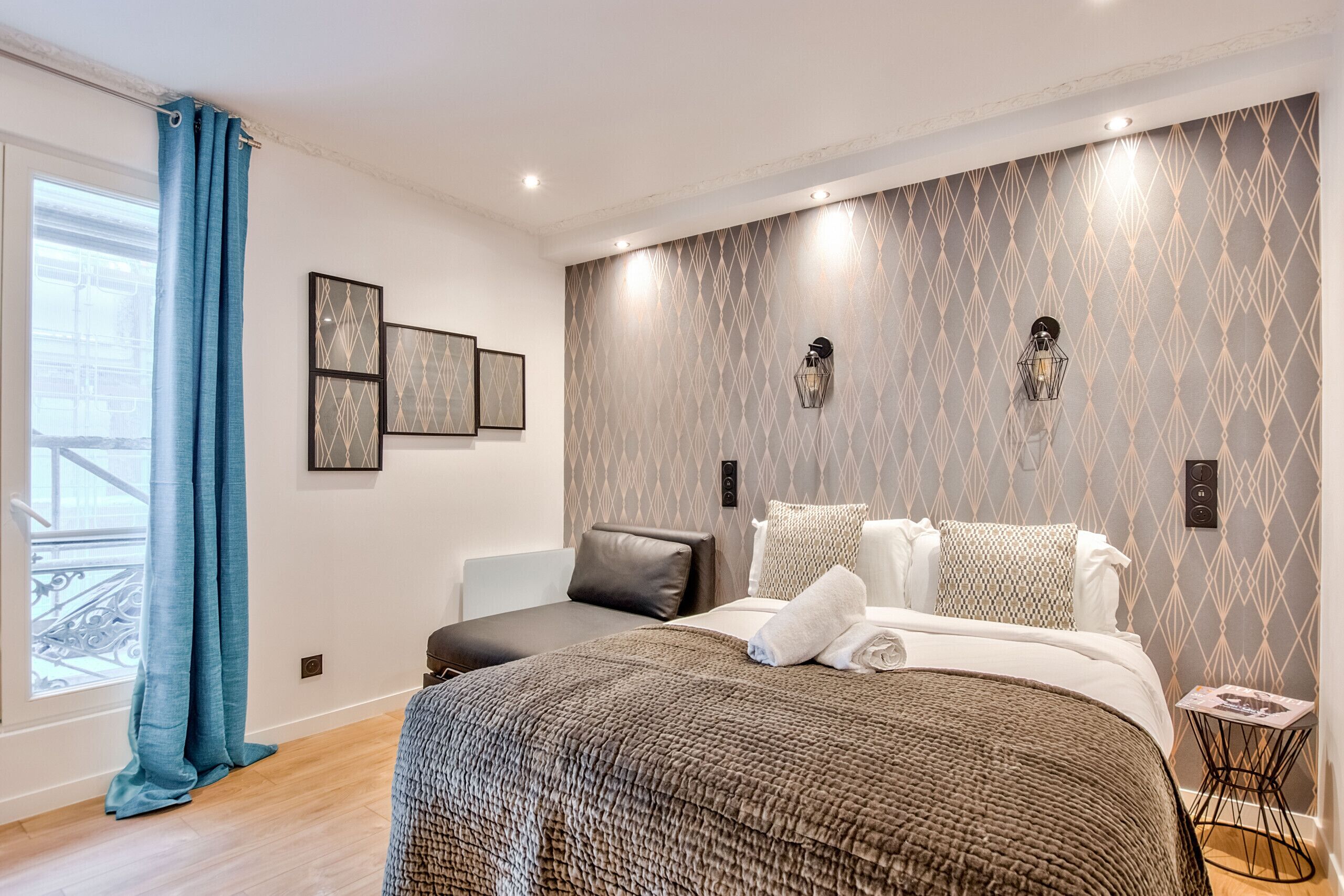 A modern bedroom with a large bed, a small seating area, and a view through a window with blue curtains.