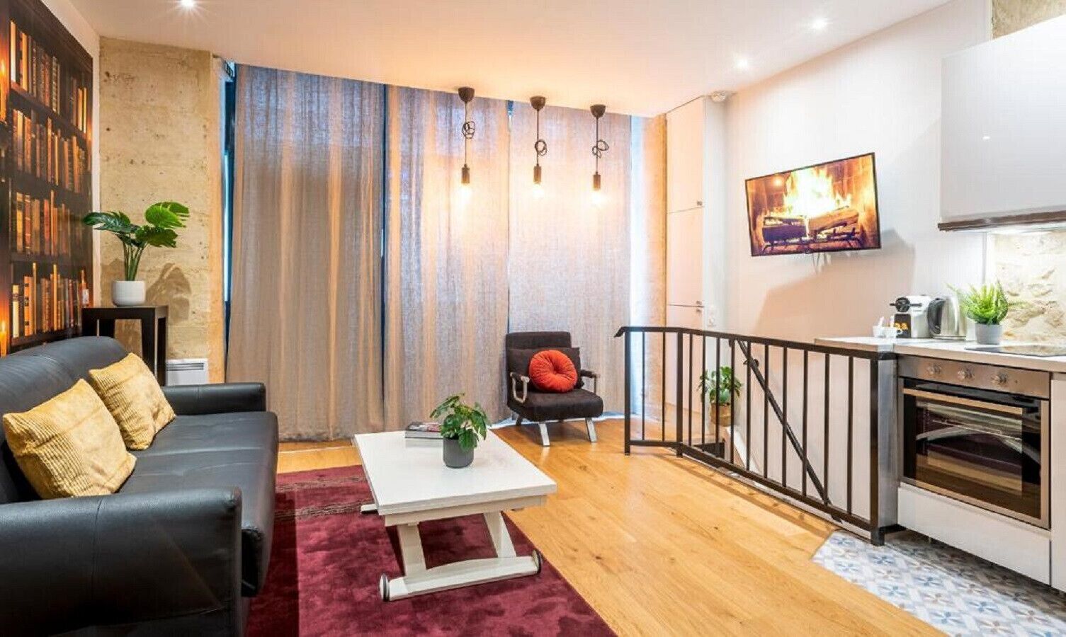 A modern living room with a black leather sofa, a white coffee table, and a red chair. There is a kitchen area with a stove and a TV mounted on the wall.