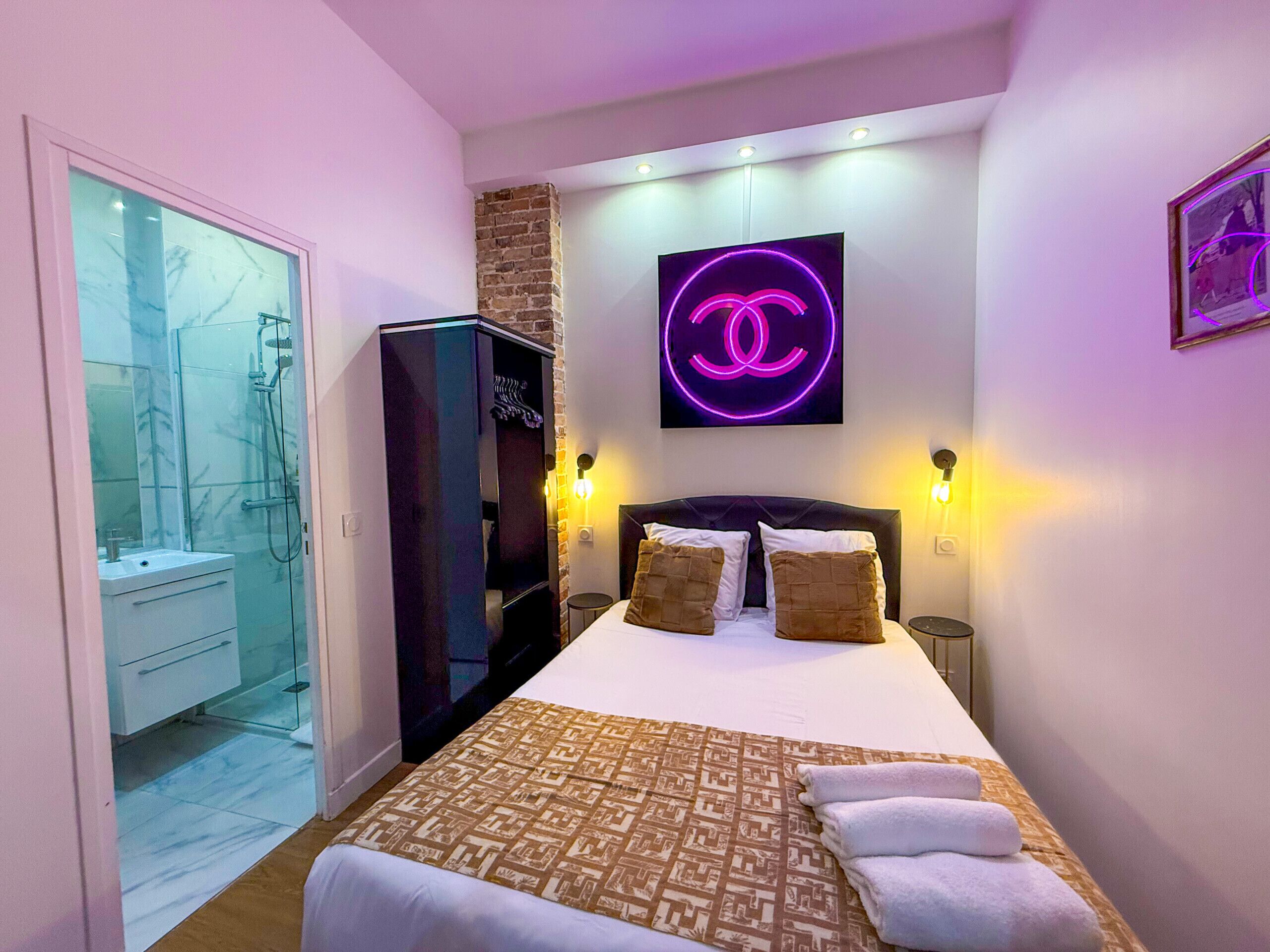 A hotel room with a bed, a wardrobe, a shower, and a neon sign.