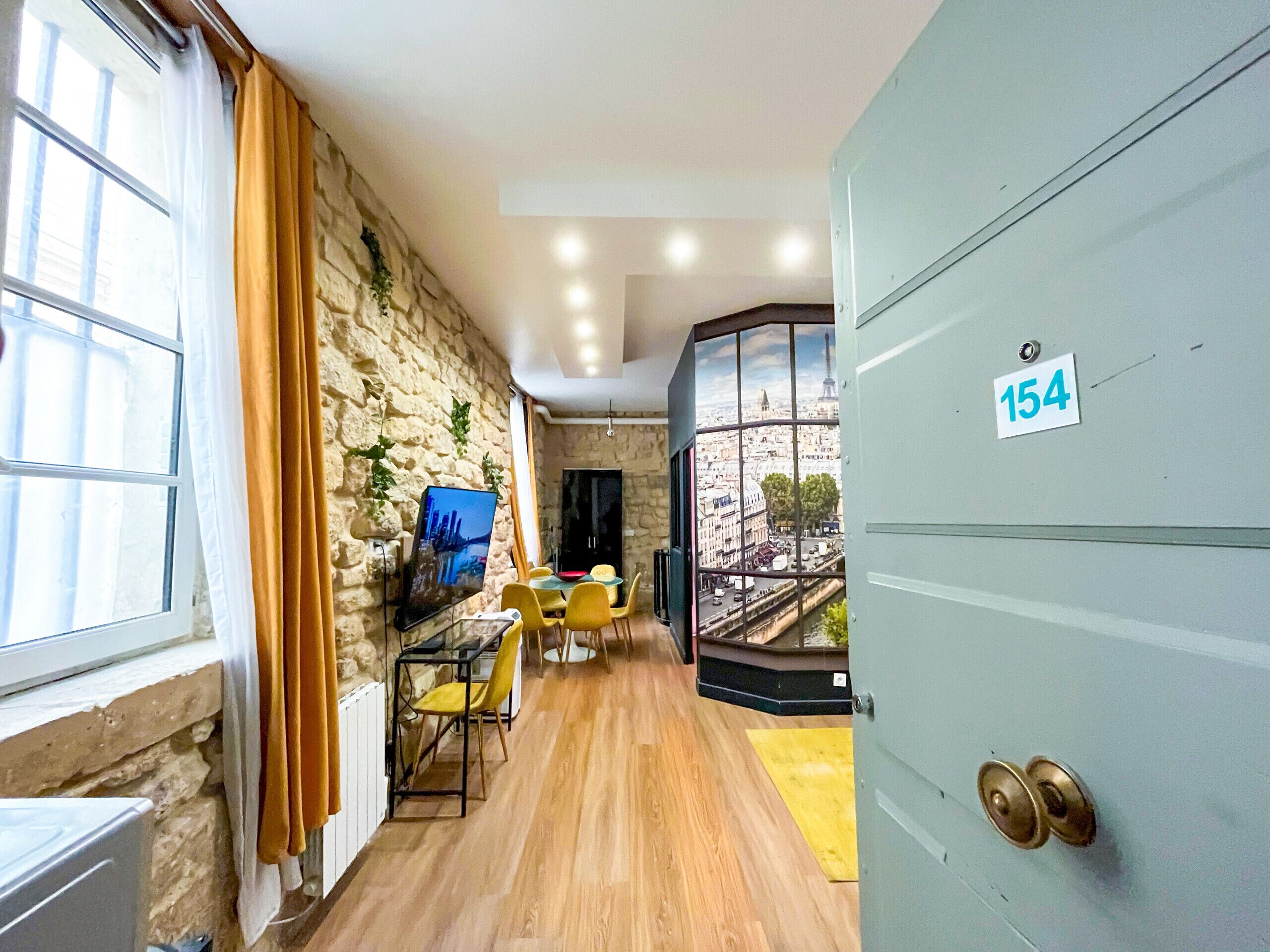 A room with a stone wall, a view of the Eiffel Tower, a dining area with yellow chairs, and a door labeled 154.
