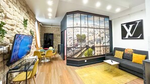 A modern room with a large window offering a cityscape view, a black sofa with yellow cushions, a glass table, and a stone wall. - Urban Flat 154 - Pretty 2 BDR in center of Paris (París)