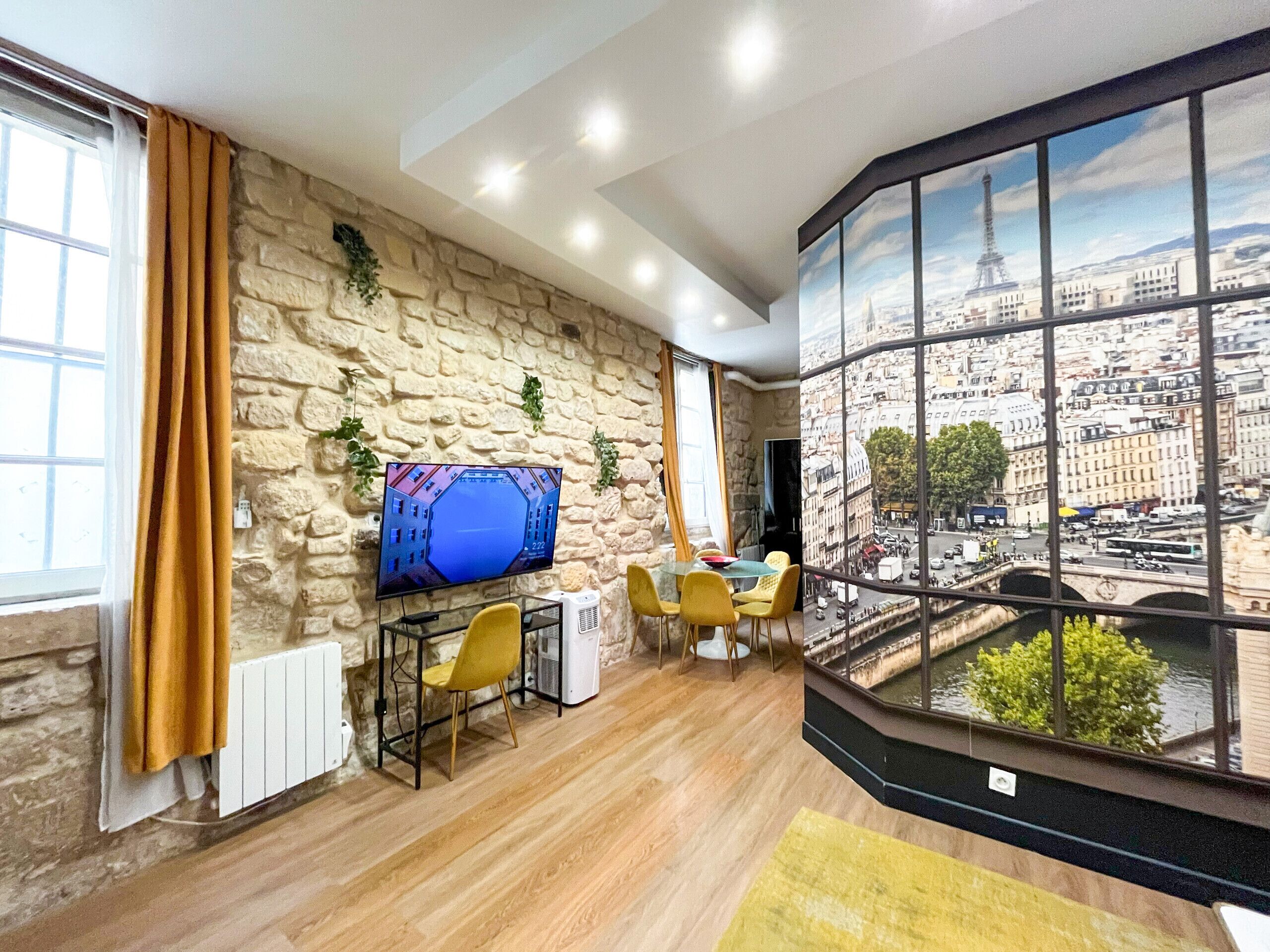 Un salon moderne avec un mur en pierre, un téléviseur à écran plat, un coin repas avec des chaises jaunes et une grande fenêtre donnant sur le paysage urbain.