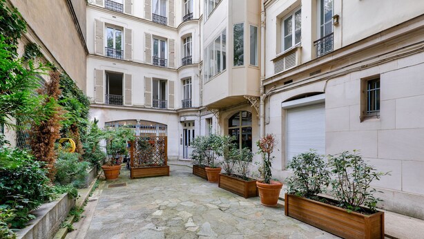 A courtyard with stone flooring, potted plants, and a building with multiple balconies and windows.