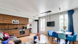 A modern kitchen with wooden cabinets, a built-in oven, and a microwave. There is a dining area with blue chairs and a small table.