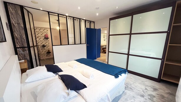 A modern bedroom with a large bed, a wardrobe, and a bathroom visible through a glass door. - Urban Flat 166 - 4BDR Luxury Apartment in Center of Paris (París)