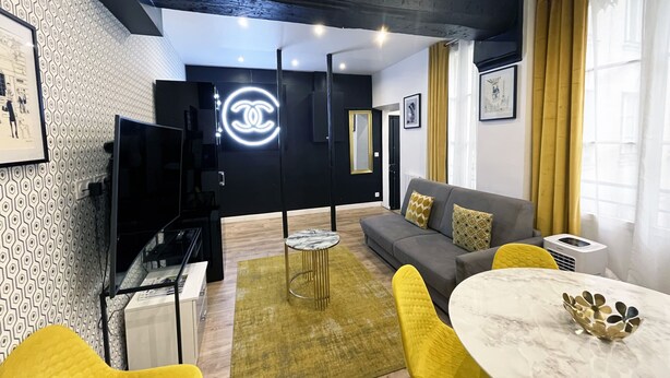 A modern living room with a grey sofa, yellow chairs, a marble table, and a Chanel logo on the wall. - Urban Flat 157 - Flat by Coco in Montorgueil (París)
