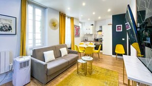 Ein modernes Wohnzimmer mit grauem Sofa, gelben Sesseln, einem Esstisch und einer Küchenzeile im Hintergrund.
