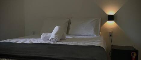 A neatly made bed with a dark-colored blanket, white pillows, and a folded towel on the bed. There is a wall-mounted light fixture on the right side of the bed.