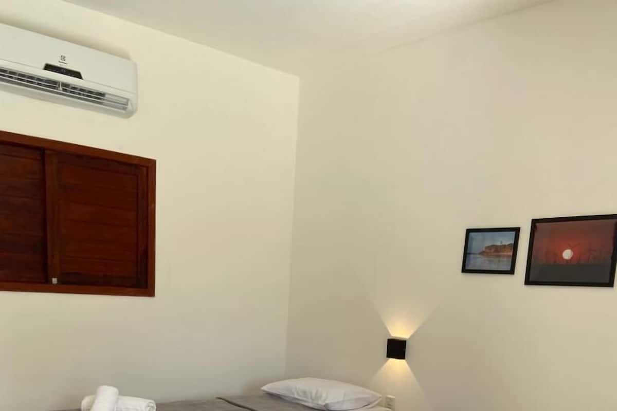 Ein Hotelzimmer mit zwei Betten, jeweils mit grauer Bettdecke und weißen Kissen. Über dem linken Bett ist eine Wandklimaanlage angebracht, und zur Rechten hängen zwei gerahmte Bilder an der Wand.