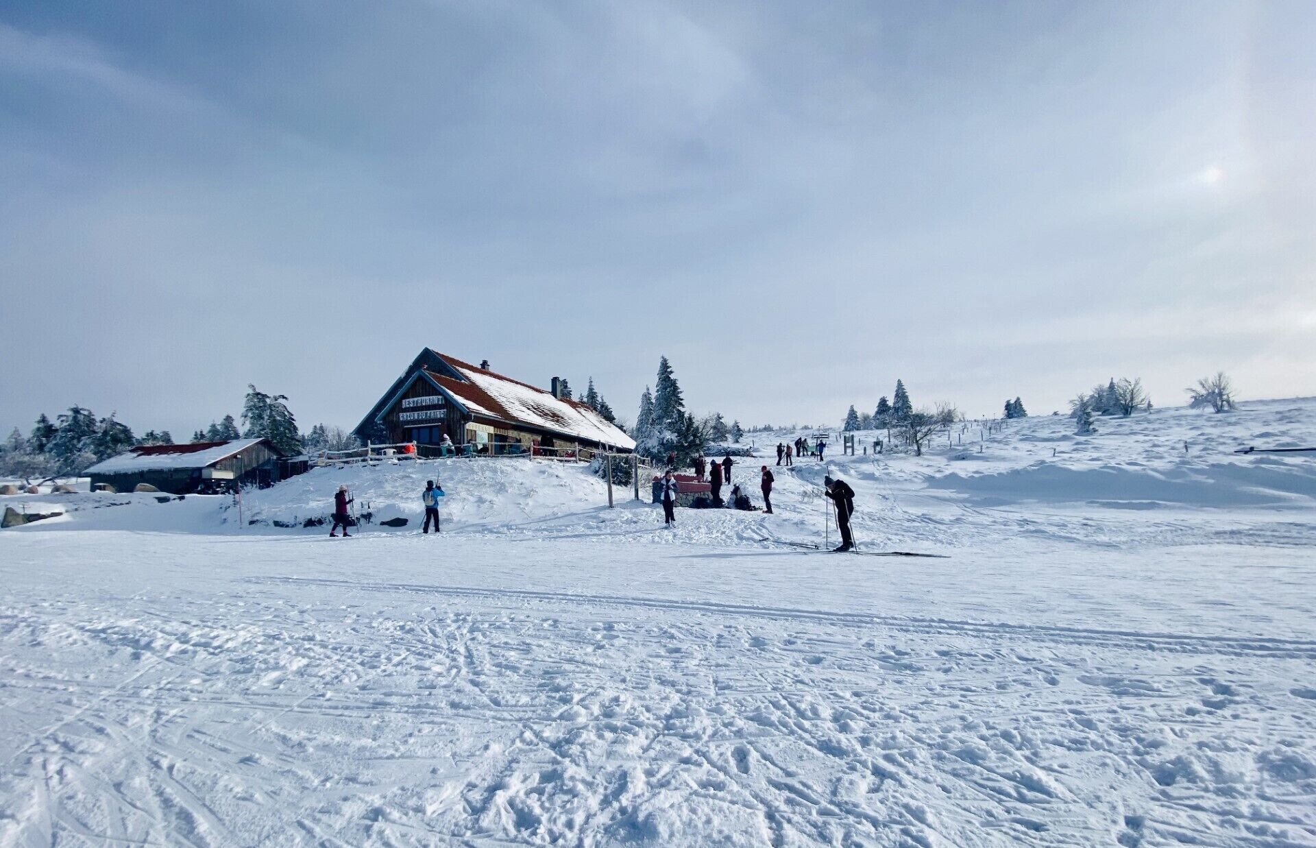A rustic wooden building with a sloped roof, likely a restaurant or lodge, surrounded by snow-covered trees and a ski slope.