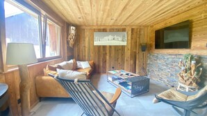 A cozy cabin-style room with wooden walls and ceiling, a sofa, a chair, a coffee table, and a TV mounted on the wall. - Le P'tit Curieux - Rupt-sur-moselle (Rupt-sur-Moselle)