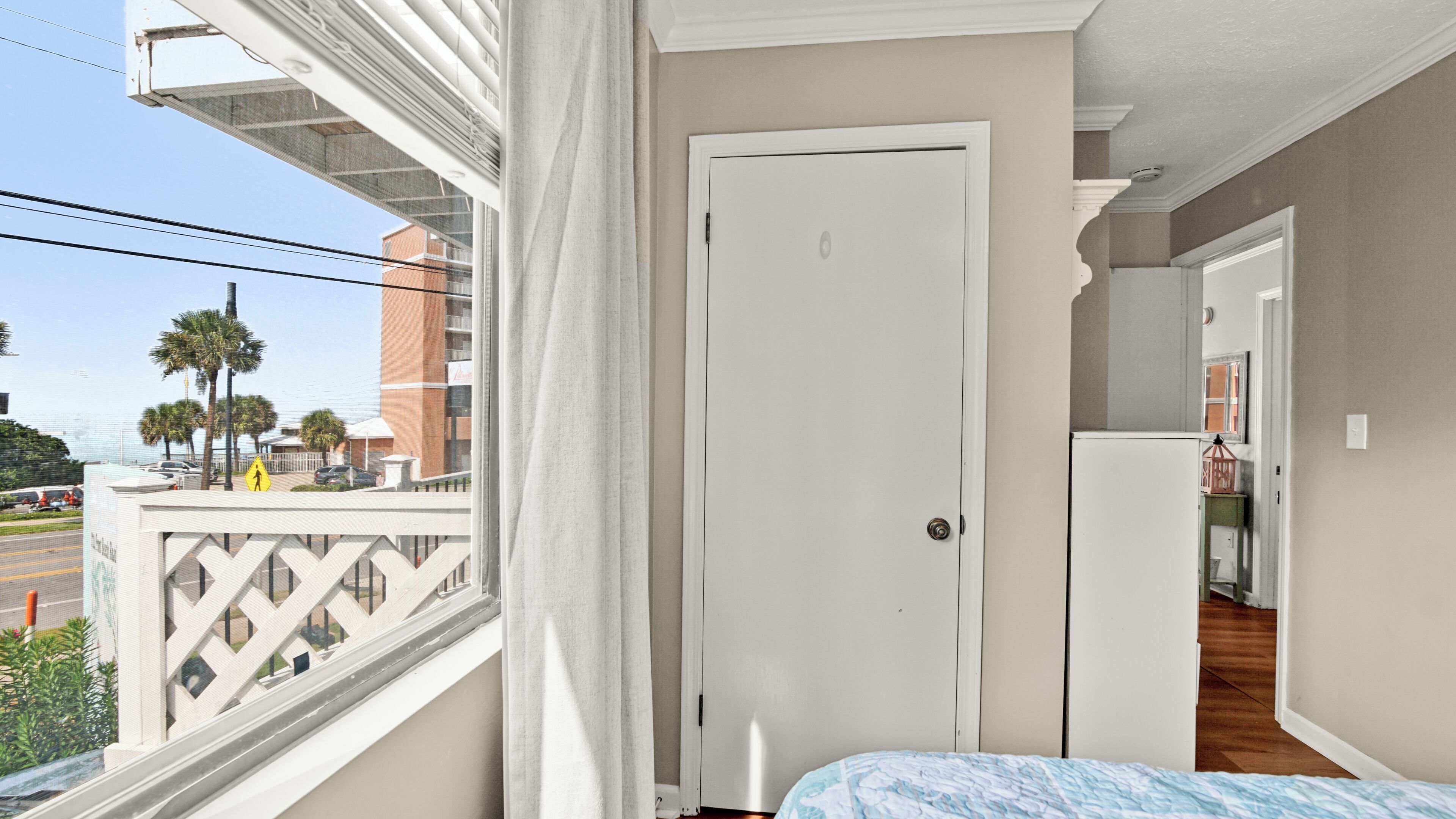 A balcony with a view of palm trees and a building, a white door, and a bed with a patterned bedspread.