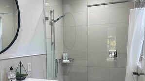 A modern bathroom with a glass shower enclosure, a white toilet, and a vanity with a mirror and toiletries. - Charming Studio Prime Location Steps from Beach (Fort Lauderdale)