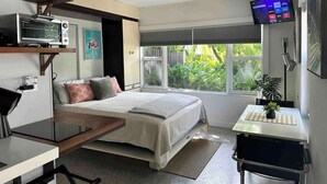A modern bedroom with a bed, a desk, a TV, and a window with a view of greenery. - Charming Studio Prime Location Steps from Beach (Fort Lauderdale)