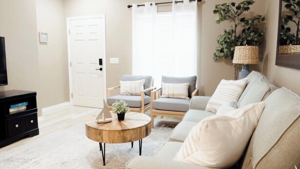A living room with a beige sofa, two armchairs, a coffee table, a TV stand, and a door.