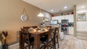 A dining area with a wooden table set for four, a clock on the wall, and a kitchen in the background with modern appliances.