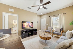 A living room with a television, a ceiling fan, a sofa set, a coffee table, and a window with curtains.