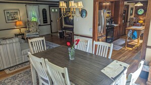 A dining area with a wooden table and white chairs, a chandelier, and a kitchen in the background.