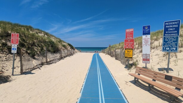 A beach path with blue railings, signs indicating parking rules, and a wooden bench.