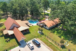 Una casa grande con piscina, dos coches aparcados en la entrada y un patio cercado.