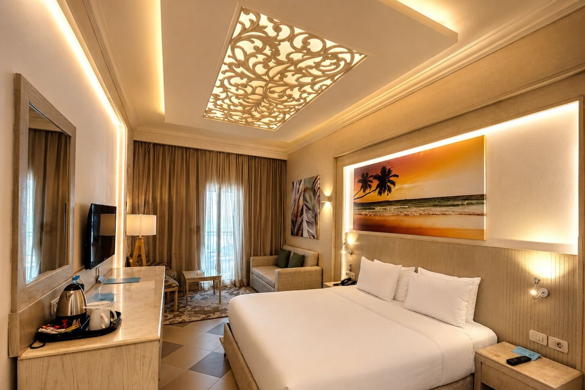 A hotel room with a large bed, a desk with a coffee maker, a TV, and a painting of a beach scene.