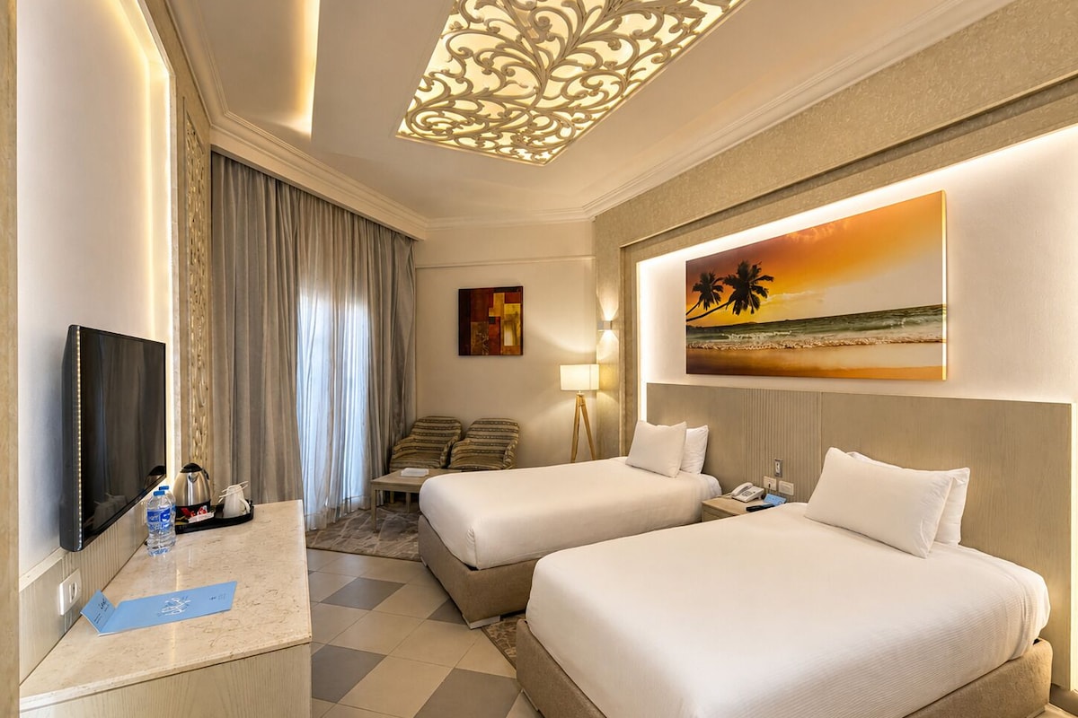 A hotel room with two beds, a desk with a flat-screen TV, a lamp, and a painting of a beach scene.