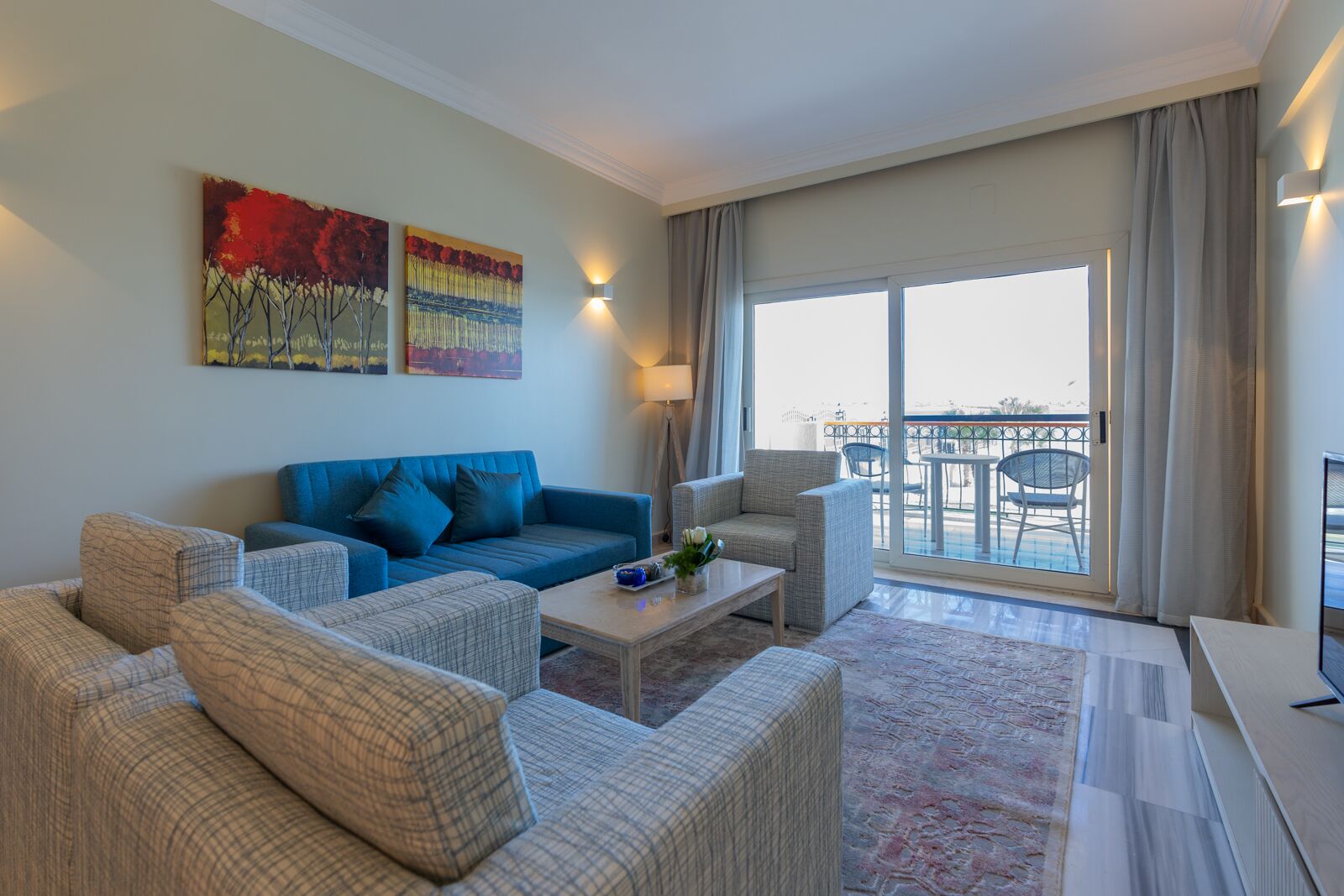 Een moderne woonkamer met een blauwe bank, een grijze fauteuil en een salontafel. Twee schilderijen hangen aan de muur, een balkon met een tafel en stoelen, en een groot raam met gordijnen.
