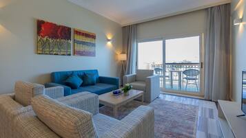 Un salon moderne avec un canapé bleu, un fauteuil gris et une table basse. Deux tableaux ornent le mur, il y a un balcon avec une table et des chaises, ainsi qu’une grande fenêtre avec des rideaux.