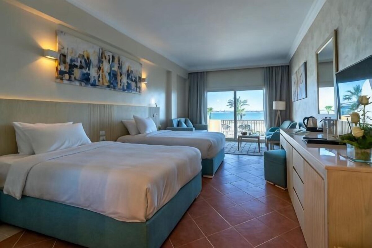 A hotel room with two beds, a desk, a chair, and a view of the sea.