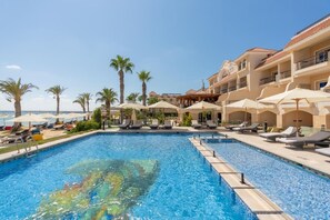 Una piscina con sillas de descanso y sombrillas, rodeada de palmeras y un edificio con balcones.
