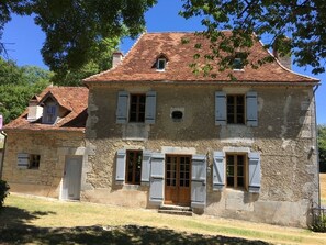 Un bâtiment en pierre avec un toit en tuiles, de nombreuses fenêtres dotées de volets et une porte d’entrée en bois située au centre.