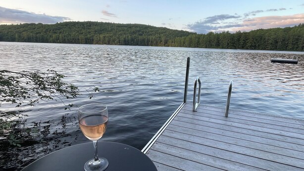A wooden dock extending into a calm lake with a glass of wine on a round table.