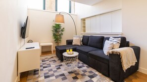 Een moderne woonkamer met een bank, een klein bijzettafeltje, een aan de muur bevestigde televisie, een lamp en een plant.