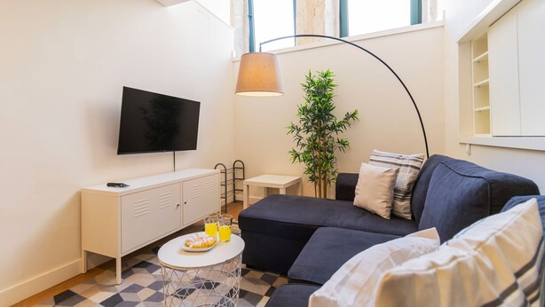 A modern living room with a flat-screen TV, a sofa, a coffee table with a plate of food and a glass of orange juice, a small white table, a tall green plant, and a lamp. - Delightful Duplex by LovelyStay (Vila Nova de Gaia)