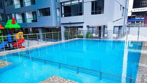 Une piscine avec une terrasse carrelée, entourée d’une clôture en métal et un bâtiment visible au fond.