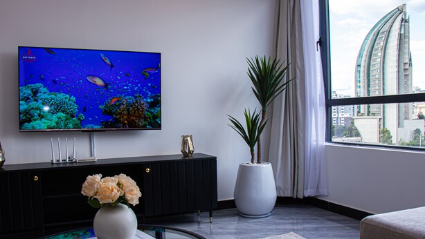 Un salon moderne avec un téléviseur à écran plat, un meuble de rangement noir, un vase blanc orné de fleurs, une table basse en verre et une grande fenêtre donnant sur la ville.