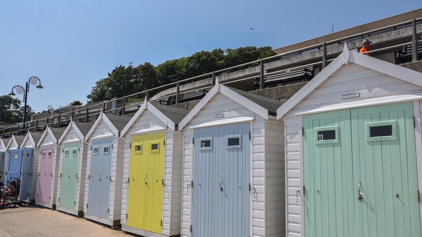 A row of colorful beach huts with numbered doors, a gravel path, and a bridge in the background. - 1 Dolphin Cottages (Lyme Regis)
