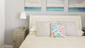 A bedroom with a bed, pillows, a nightstand, a lamp, and three wall paintings of a beach scene. - 1 Dolphin Cottages (Lyme Regis)