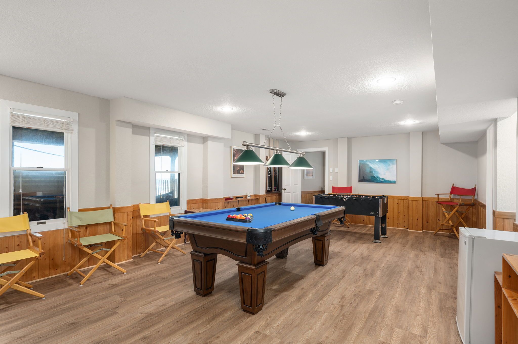 A pool table with a blue felt surface, surrounded by wooden chairs and a green overhead lamp.