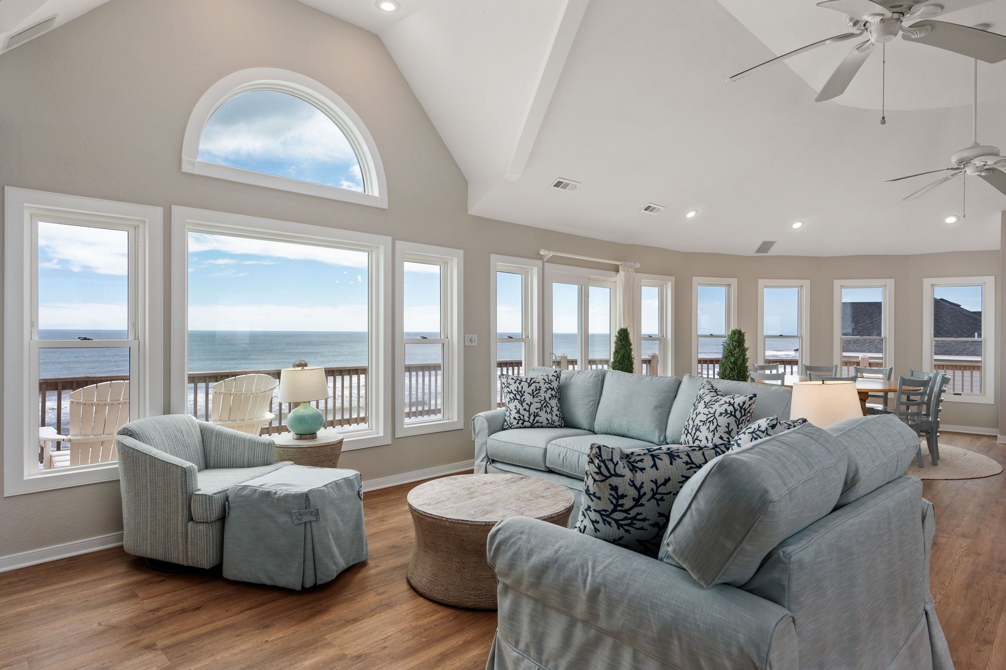 A spacious living room with a large window overlooking the ocean, a ceiling fan, and a dining area with a view.