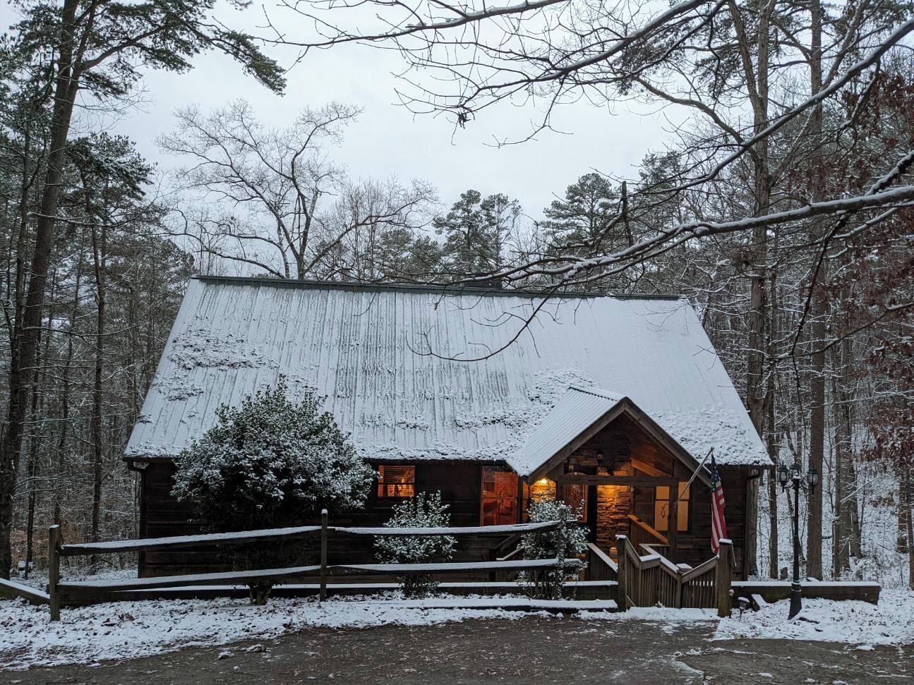 A log cabin with a snow-covered roof and a wooden porch, surrounded by trees.