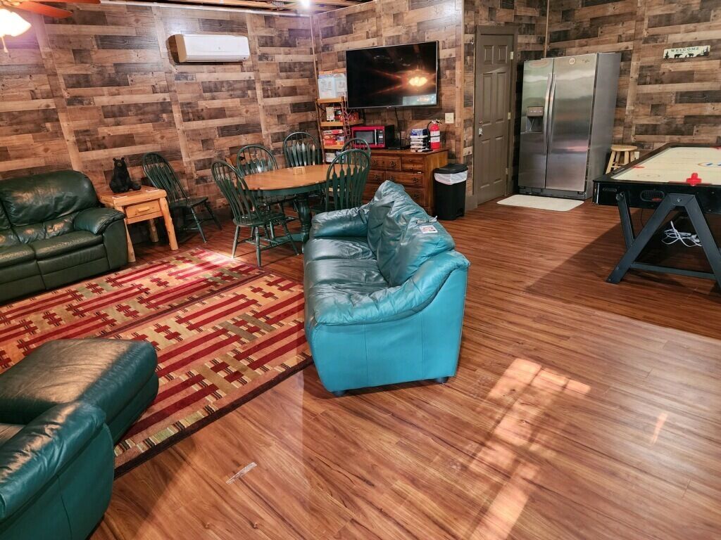 A living room with a wooden floor, a blue sofa, a green sofa, a dining table with chairs, a television, and a refrigerator.