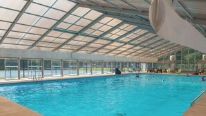 An indoor swimming pool with clear glass walls, a roof, and a poolside area with chairs and a lifeguard stand.