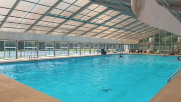 An indoor swimming pool with clear glass walls, a roof, and a poolside area with chairs and a lifeguard stand.