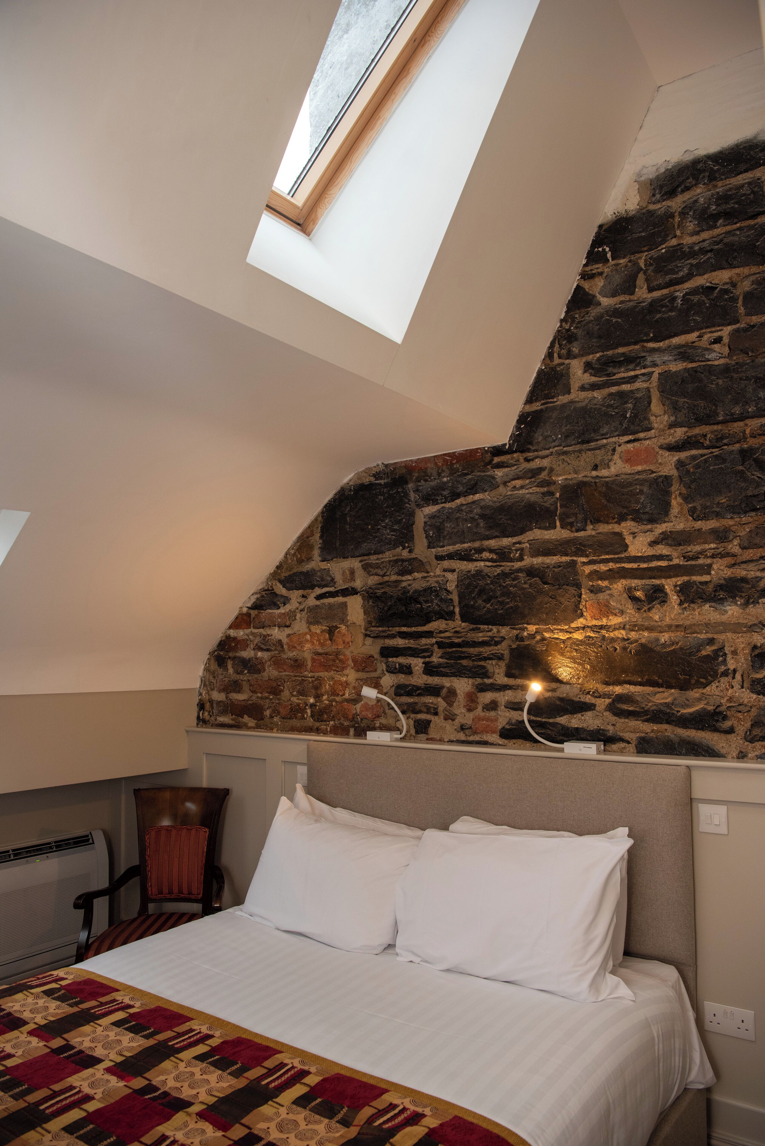 Un dormitorio con pared de piedra inclinada, tragaluces, cama con ropa de cama blanca y manta estampada.