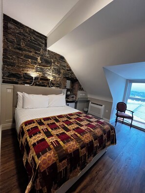 A bedroom with a large bed, a stone wall, a chair, and a view through a window.