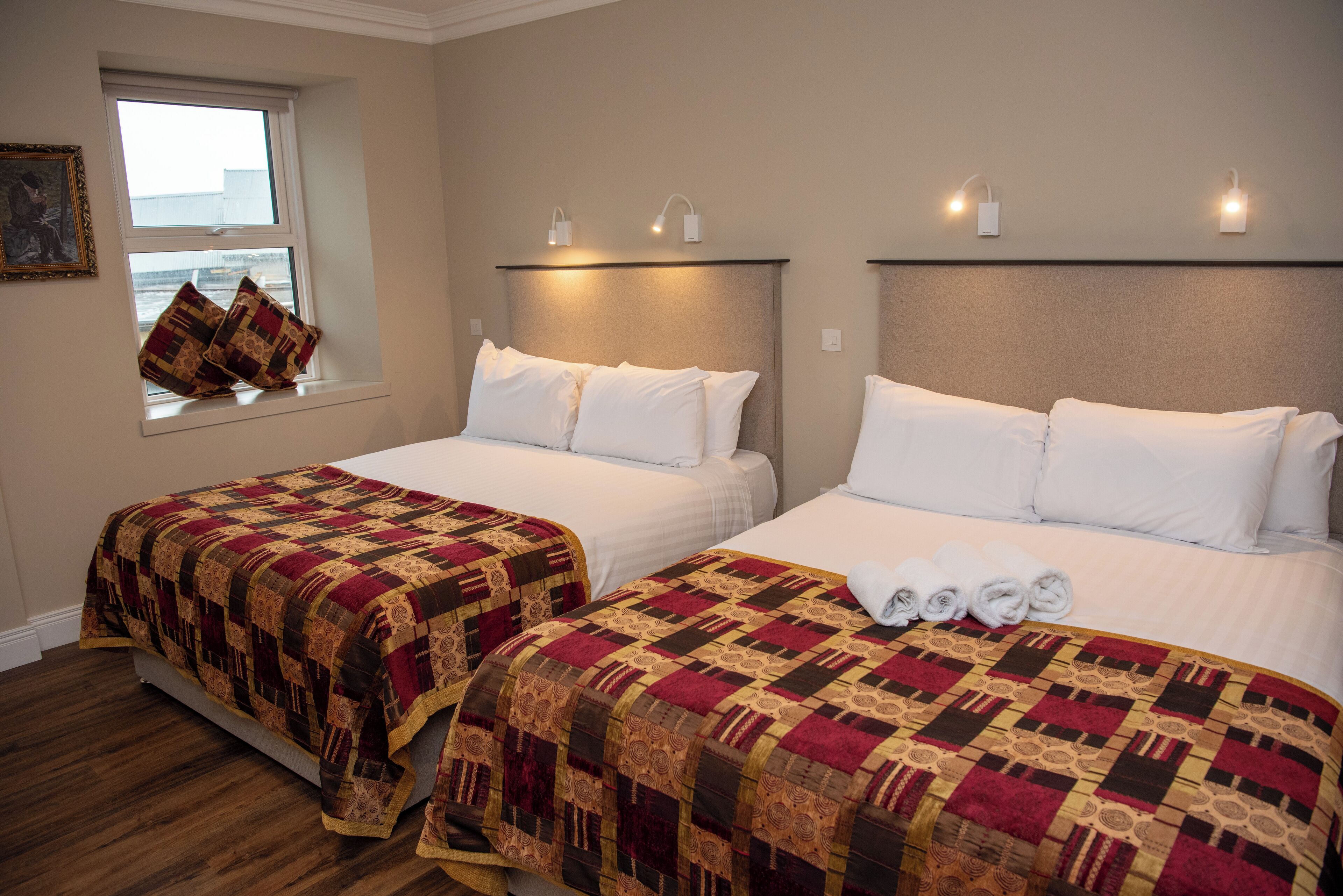 Habitación de hotel con dos camas, cada una con cubrecama acolchado, ventana con vistas y lámparas fijadas a la pared.