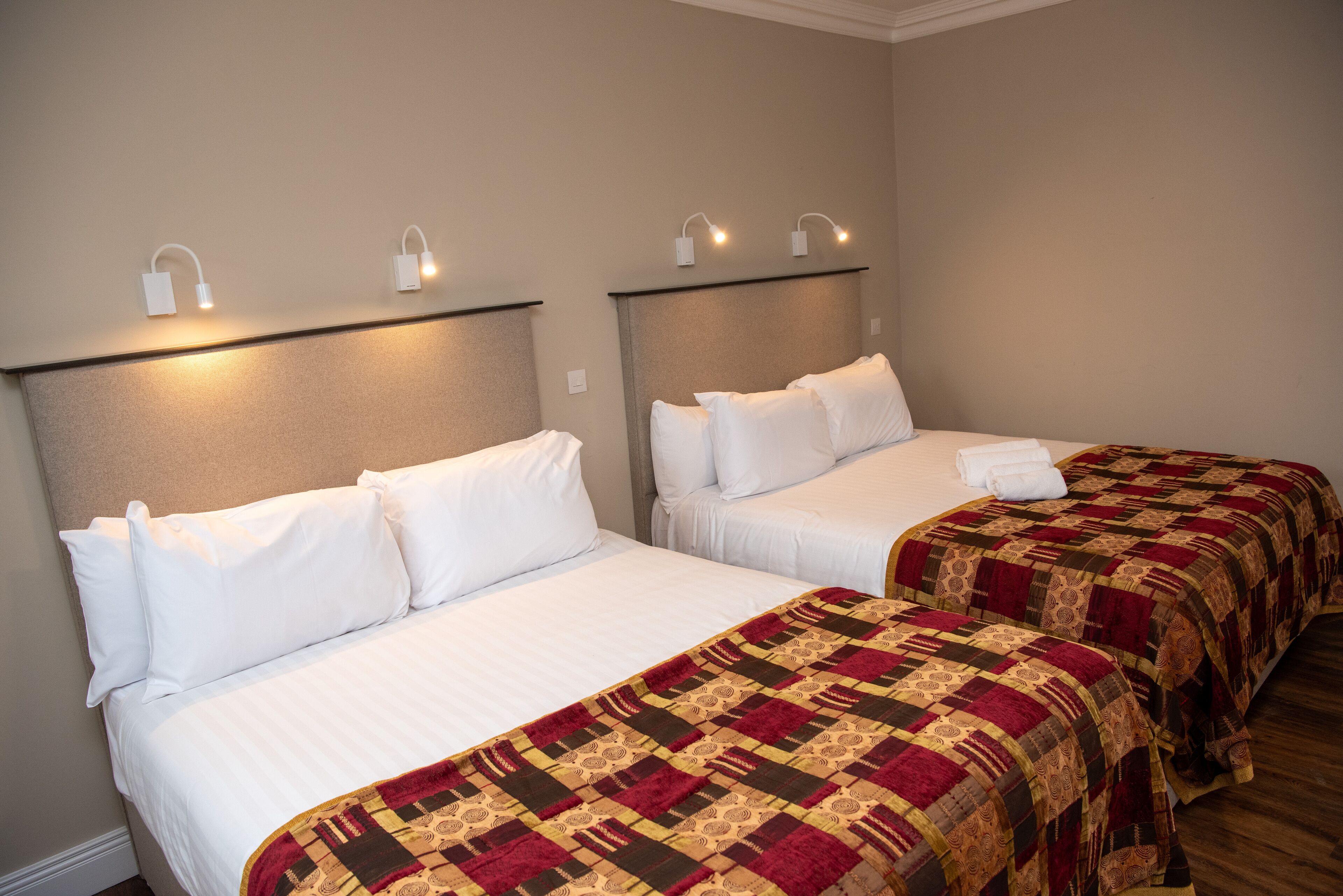 Two single beds with white linens and a patterned blanket, headboards with wall-mounted lights, and a neutral-colored wall.